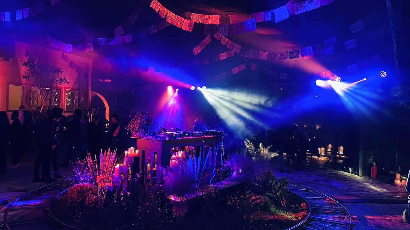 Cabina de DJ decorada con flores y velas bajo papel picado, con iluminación azul y roja en un evento de temática mexicana